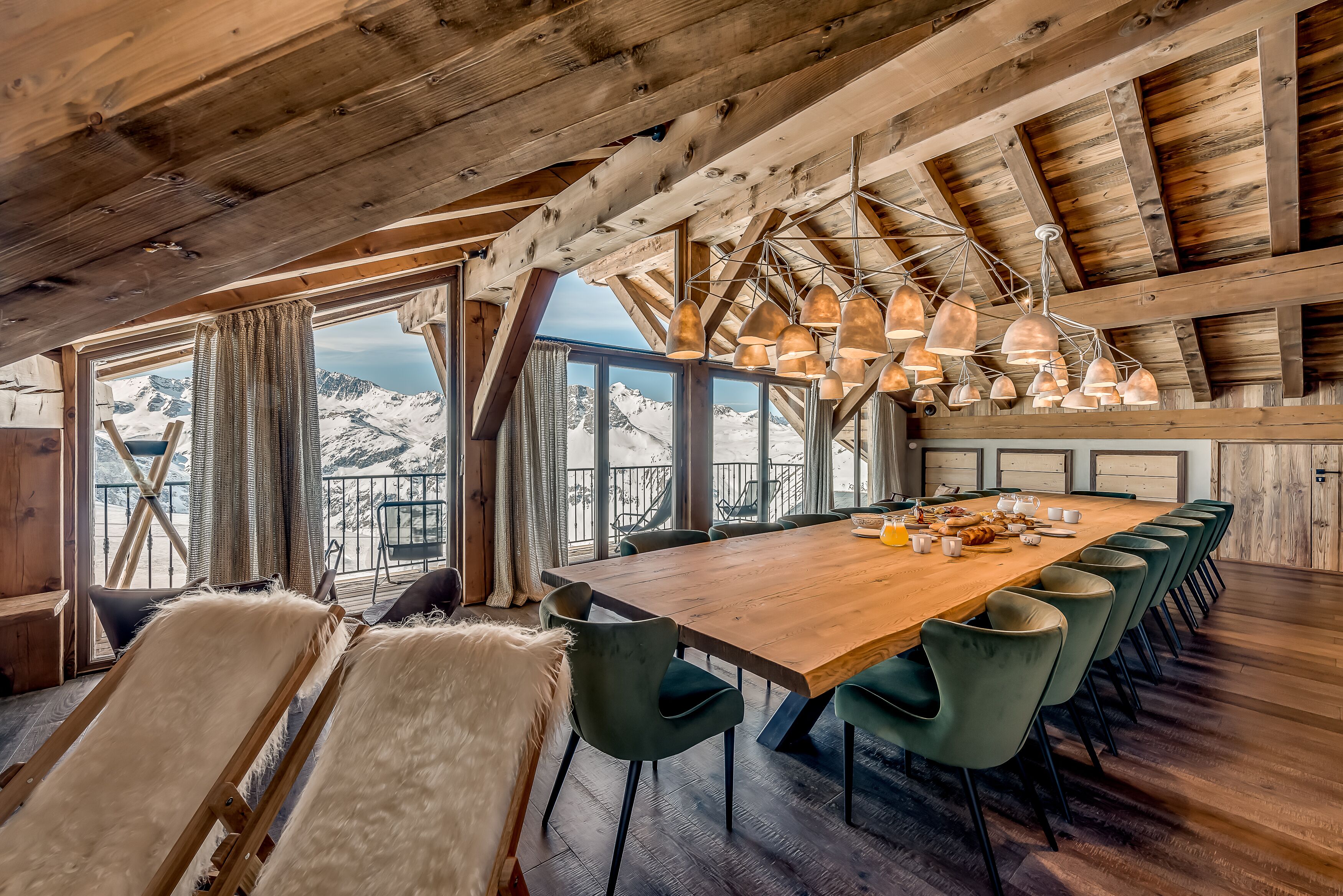 Comedor de madera con una mesa grande y sillas, ventanas amplias con vista a la montaña y lámparas colgantes.