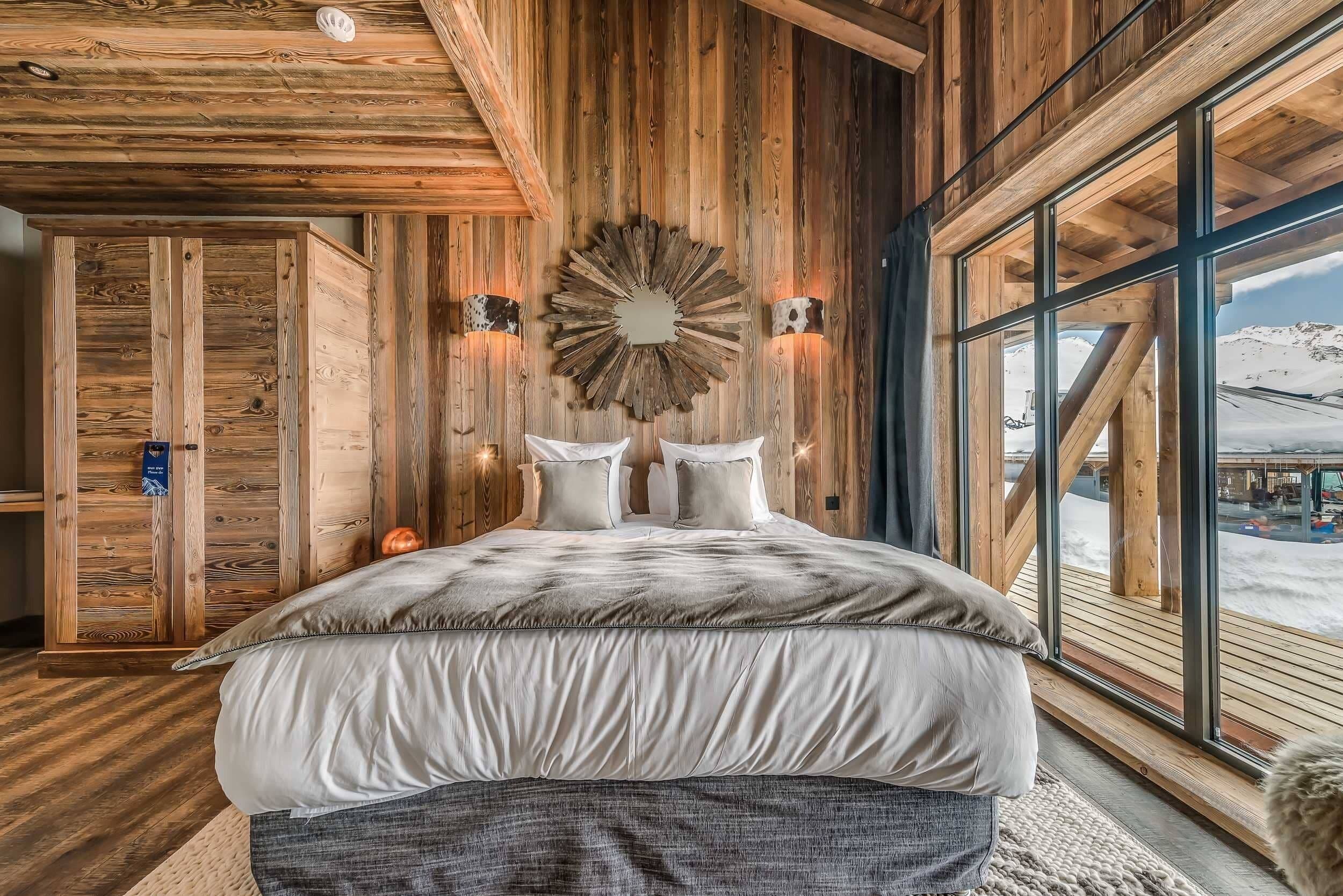 A wooden interior with a bed, large windows, and a decorative wall piece.