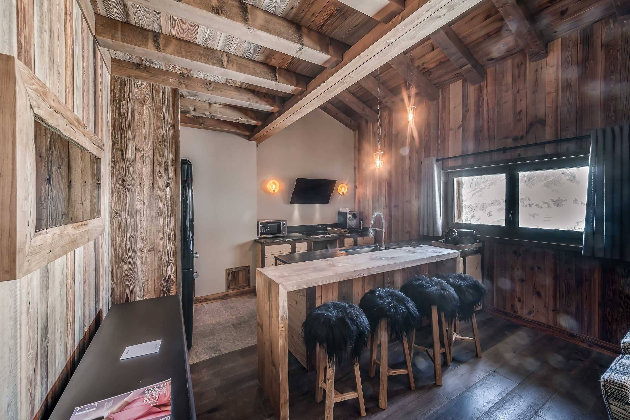 A rustic kitchen with wooden beams, a wooden island with stools, a TV mounted on the wall, and a window with a view.