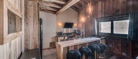 A rustic kitchen with wooden beams, a wooden island with stools, a TV mounted on the wall, and a window with a view.