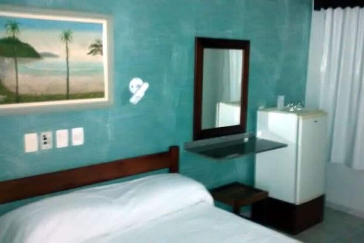 A bedroom with a bed, a small sink, a mirror, and a painting of a beach scene.