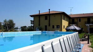 A swimming pool with a white deck, a yellow building with a red roof, and a clear blue sky. - Olistella (Palazzolo dello Stella)