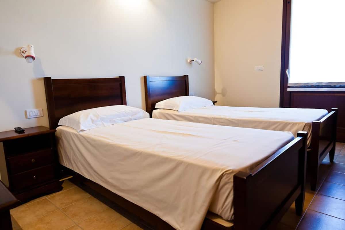 A hotel room with two single beds, a nightstand, a wall-mounted light, and a window with a view.