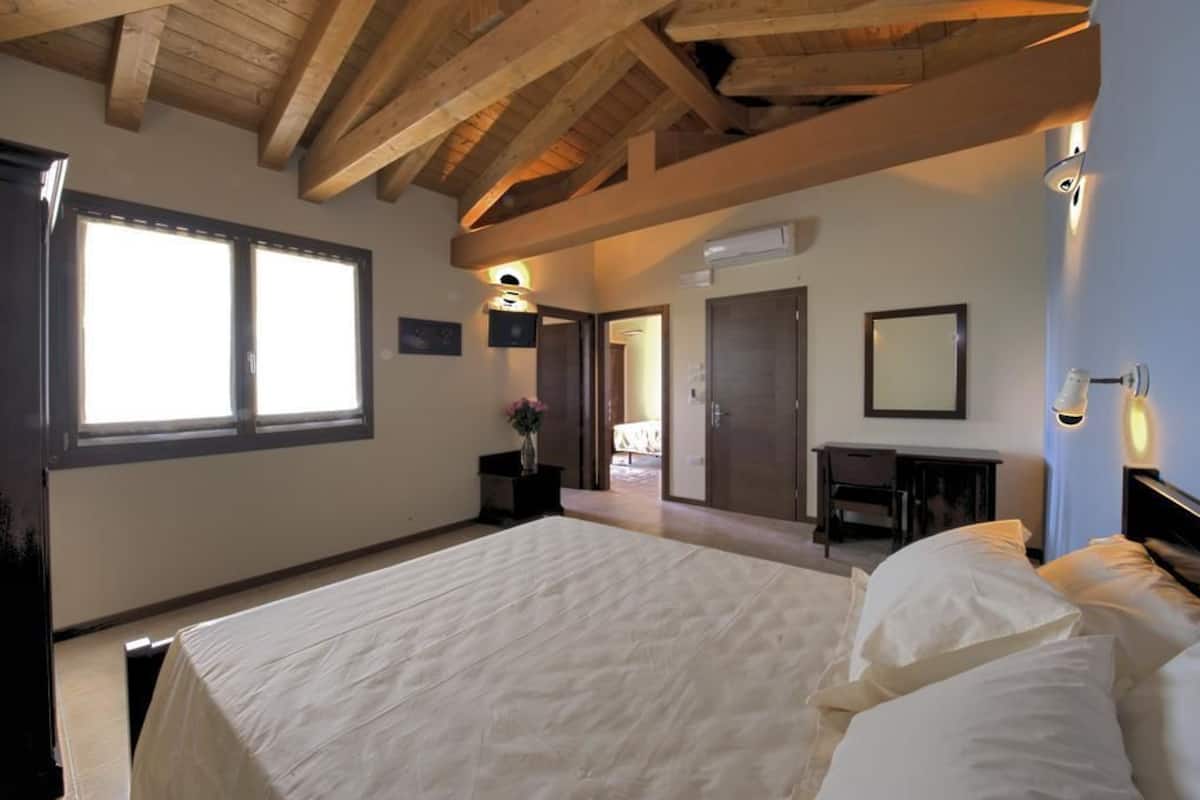 A bedroom with a large bed, wooden ceiling beams, a desk, and a chair.