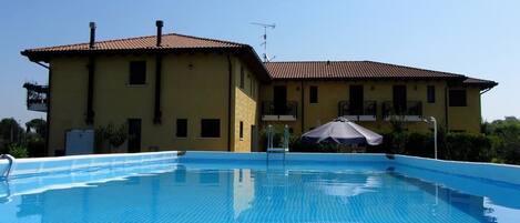 A swimming pool with a tiled edge, a yellow building with a red roof, and a white umbrella.