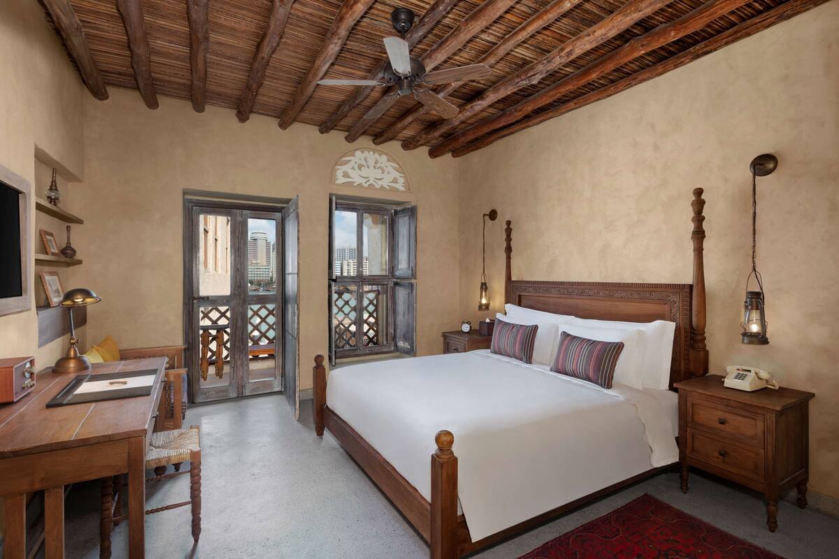Une chambre à coucher avec un plafond en bois, un lit recouvert de draps blancs, une table de chevet avec une lampe et un bureau également doté d’une lampe.