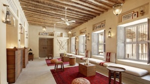 Un vaste salon doté d’un plafond haut, de poutres en bois, d’un tapis à motifs rouges et d’une grande fenêtre avec des volets blancs.