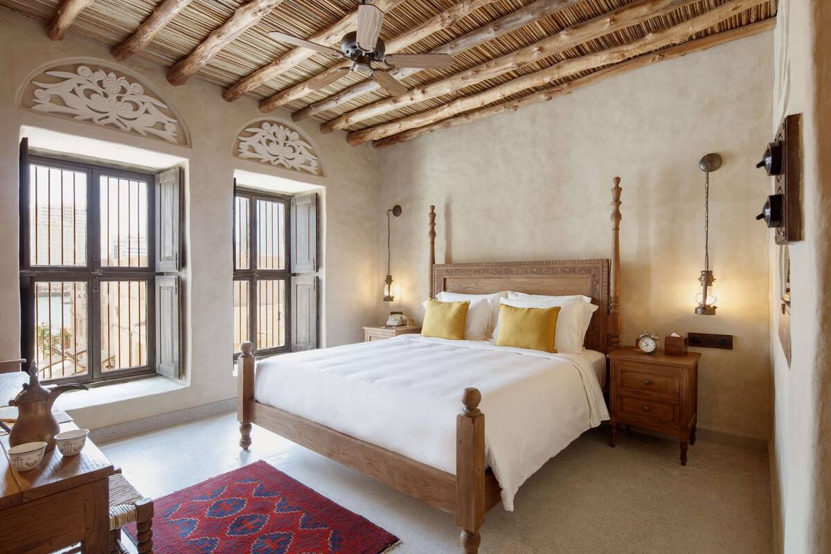 Une chambre à coucher avec un lit en bois, deux tables de chevet, une commode, un ventilateur de plafond et une grande fenêtre avec des barreaux.
