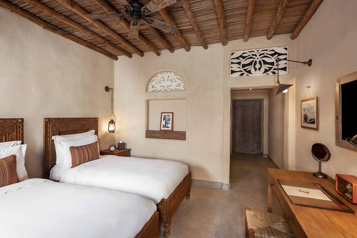 Une chambre d’hôtel avec deux lits, des têtes de lit en bois, un ventilateur de plafond, un bureau avec une chaise, un miroir et un tableau encadré au mur.