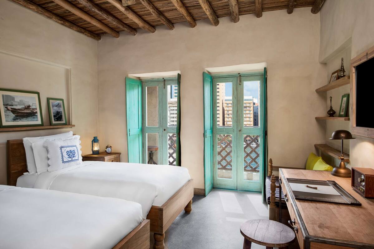 Une chambre avec deux lits, un bureau, une chaise, une lampe et une étagère ornée d’objets décoratifs.
