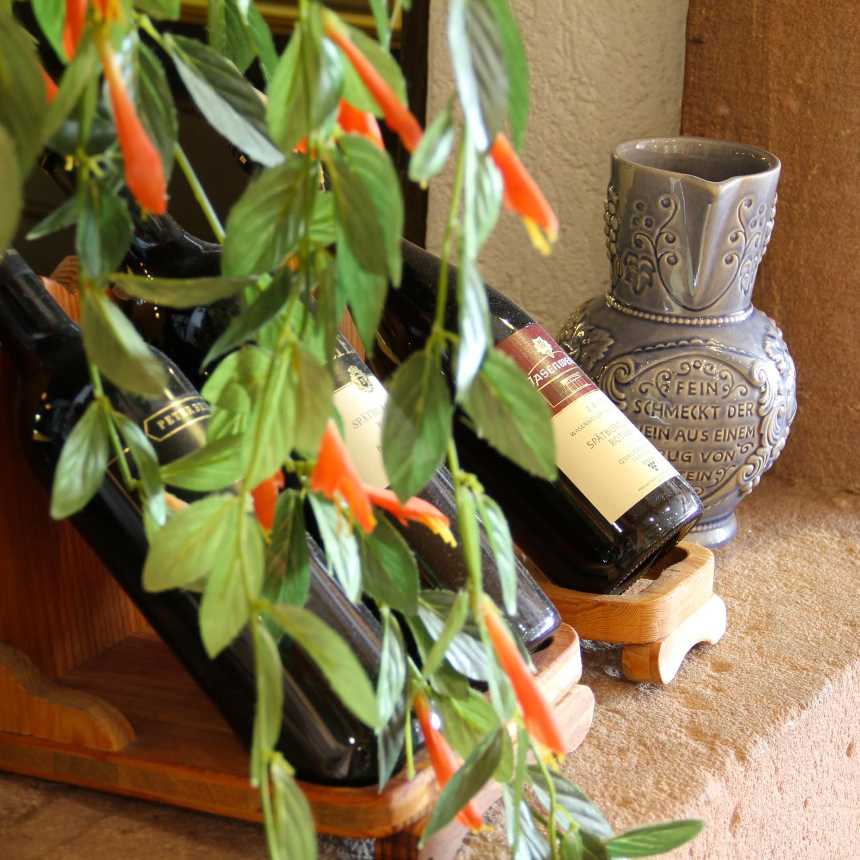 A wooden wine rack holding several bottles of wine, a decorative metal vase, and a plant with green leaves and red flowers.