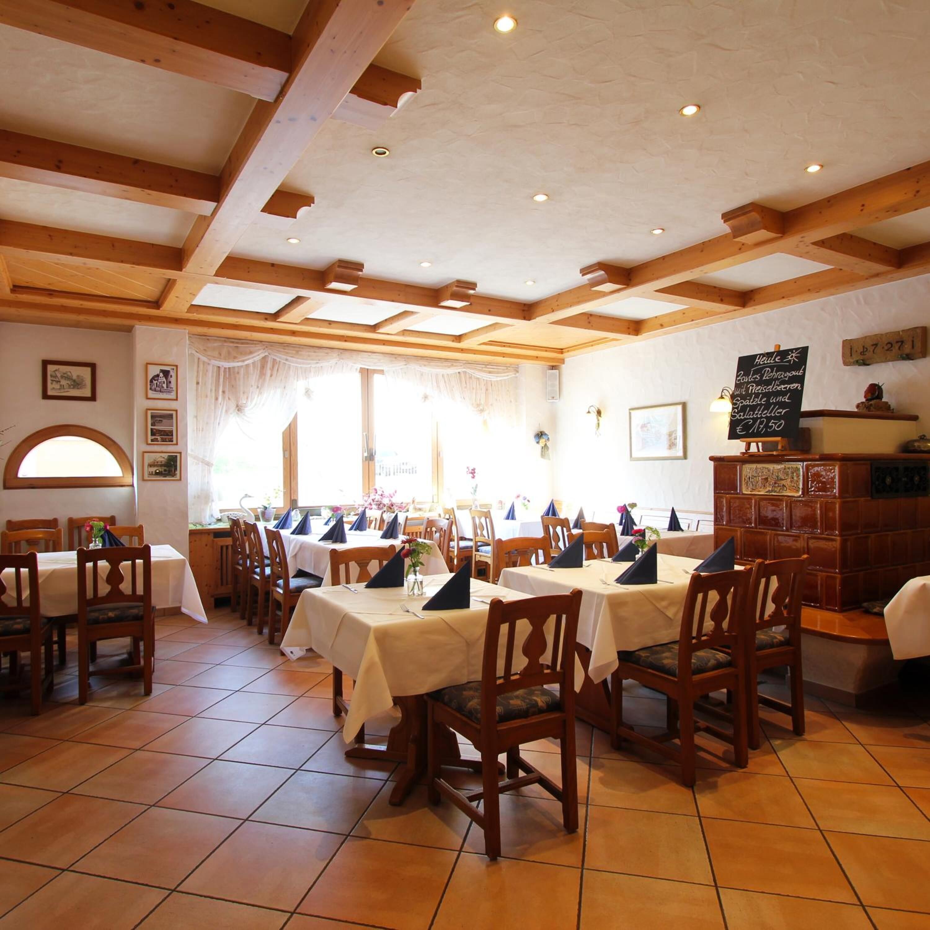A dining area with wooden beams, white walls, and tiled floors. Tables are set with chairs and napkins.