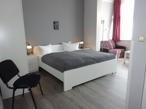 A modern bedroom with a white bed, grey walls, and a wooden floor. There is a black chair, a small table, and a lamp on the nightstand.