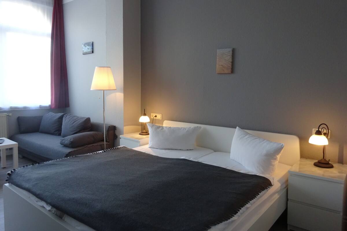 A hotel room with a bed, two bedside tables, a nightstand, a sofa, a lamp, and a window with curtains.