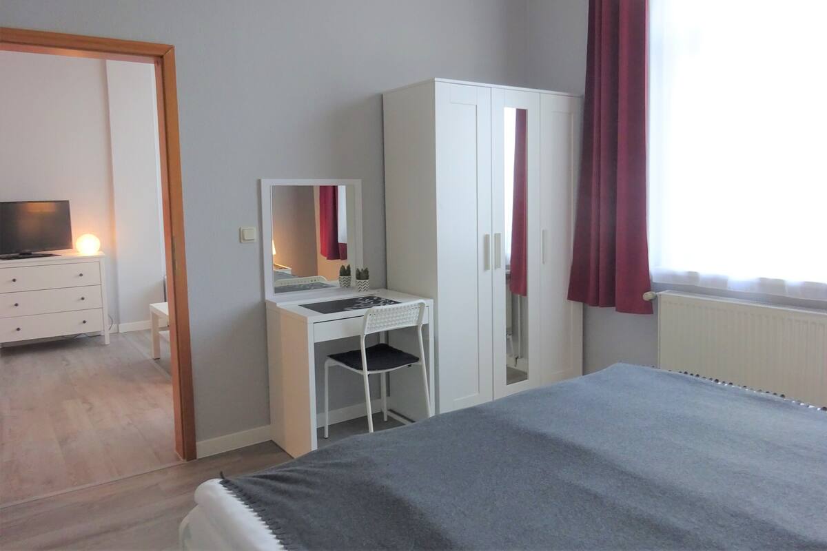 A bedroom with a bed, a desk with a mirror, a wardrobe, and a television on a dresser.