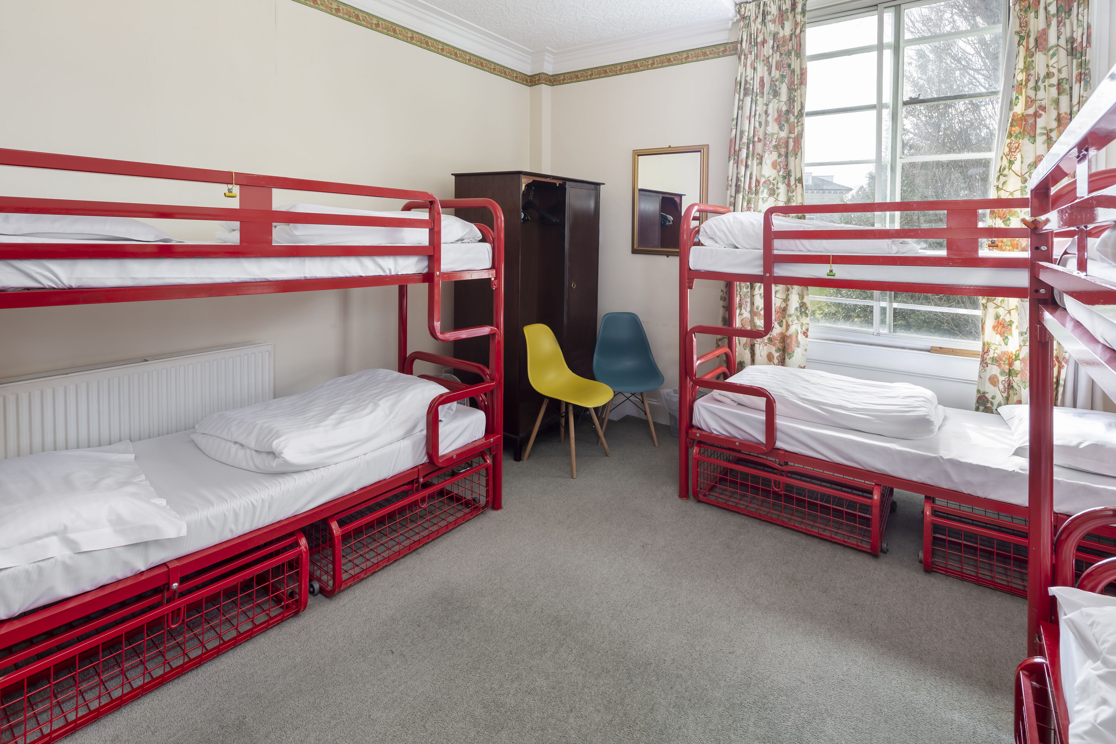 A dormitory room with bunk beds, a wardrobe, a chair, and a window with curtains.