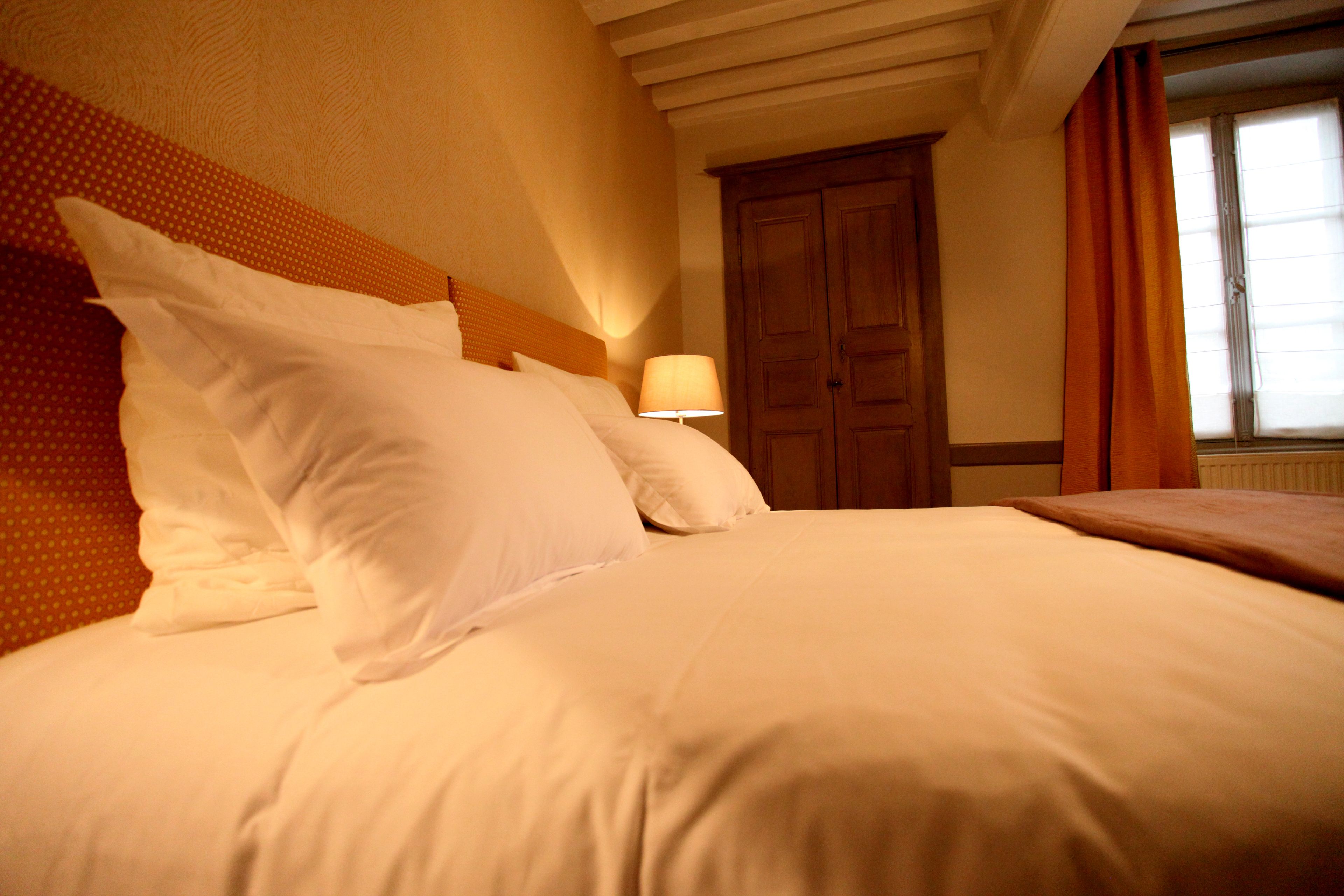 A neatly made bed with white linens and pillows, a wooden headboard, a bedside lamp, and a wooden wardrobe.