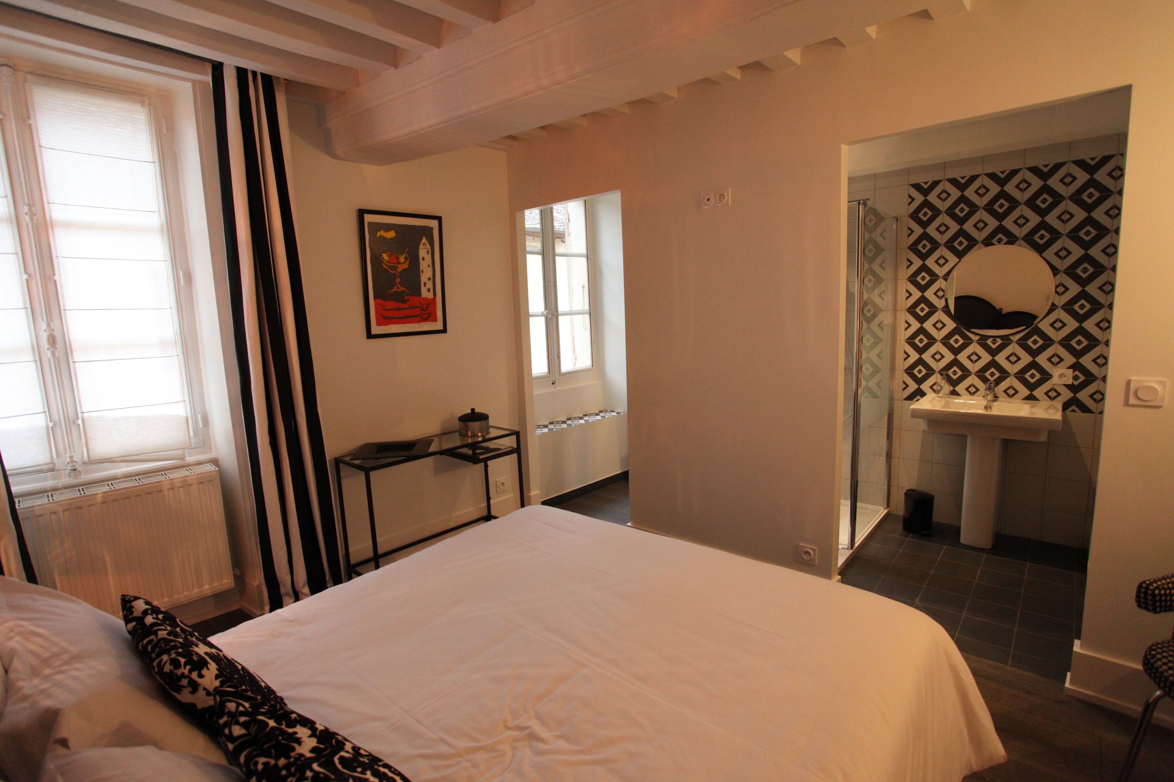 A hotel room with a bed, a small table, a window with curtains, a bathroom with a sink and mirror, and a patterned wall.