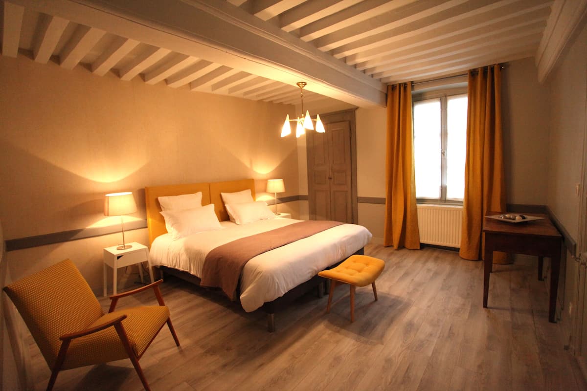 A hotel room with a large bed, a wooden chair, a small table, and a window with curtains.