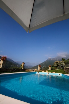 A swimming pool with clear blue water, surrounded by a stone wall and a metal fence. - Cascina del Forte (Torre Pellice)