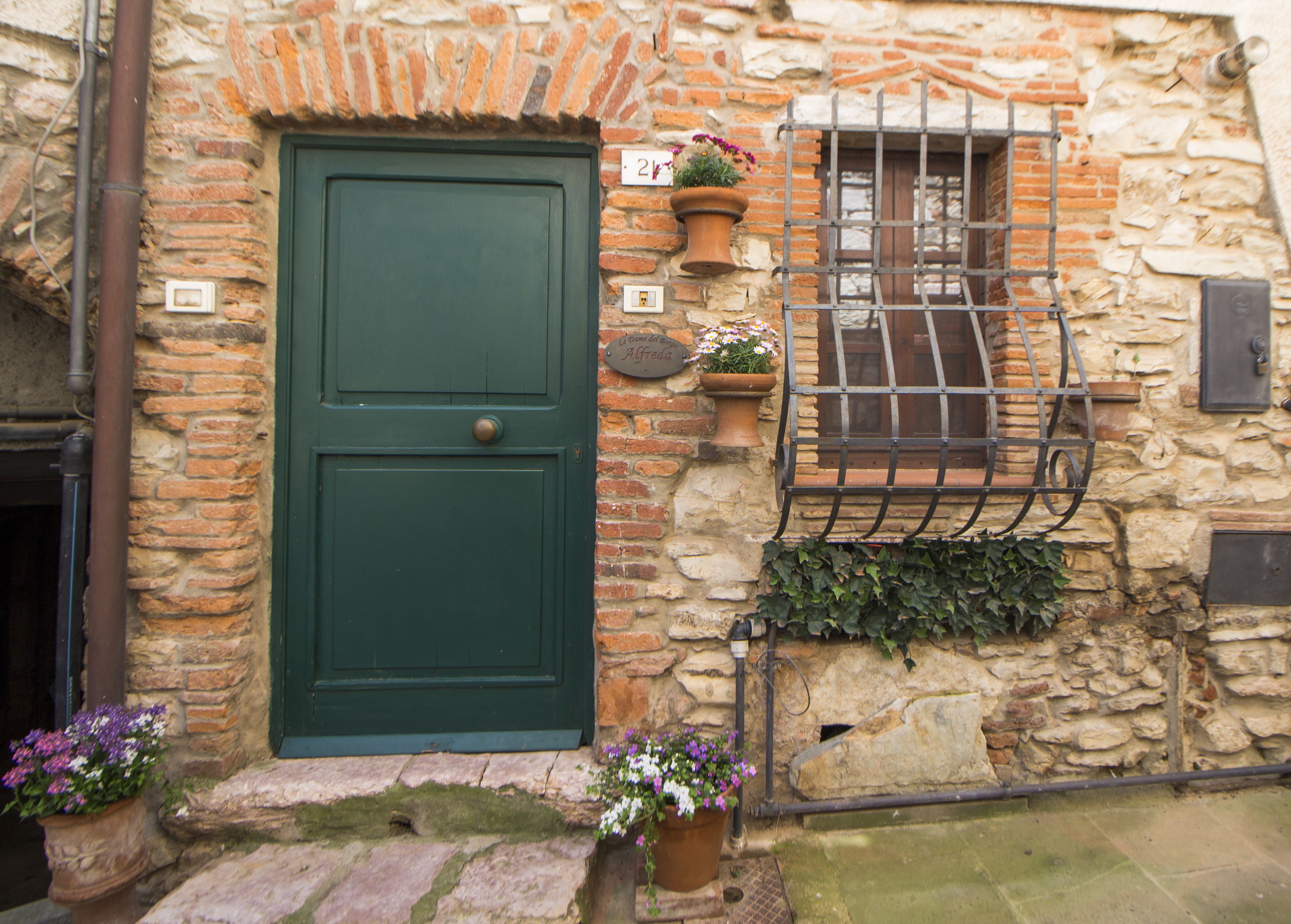 Un bâtiment en pierre avec une porte verte, une fenêtre ornée d’une grille métallique et des plantes en pot.