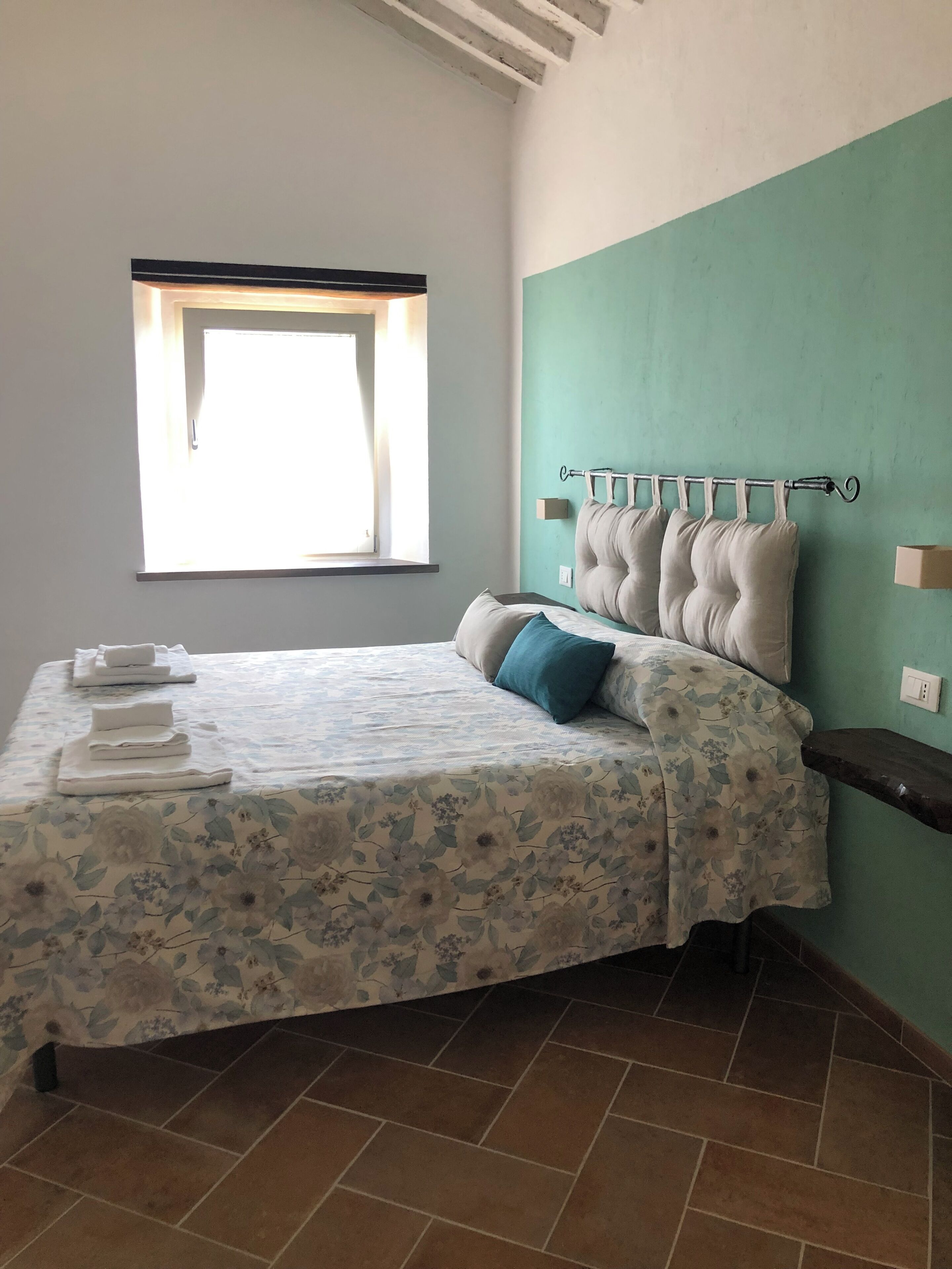 A neatly made bed with a floral bedspread, a headboard with multiple pillows, and a window with curtains.