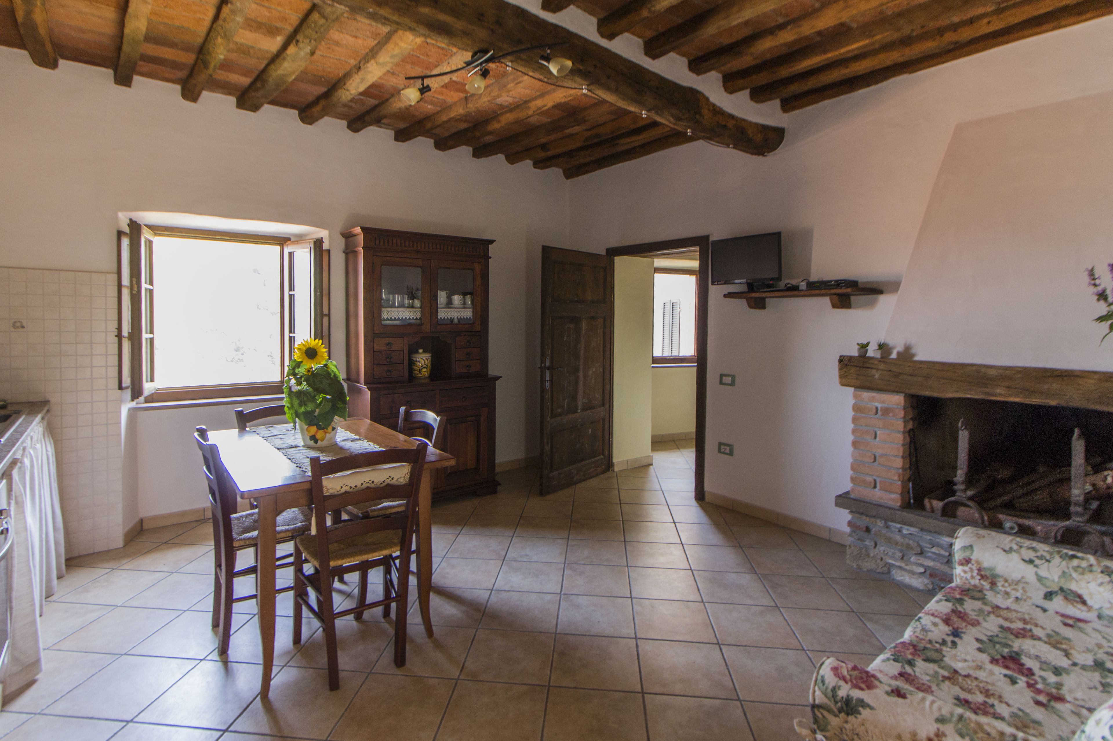 Amplia sala de estar con techo de madera, chimenea, mesa de comedor con sillas y zona de cocina con ventana y armarios.