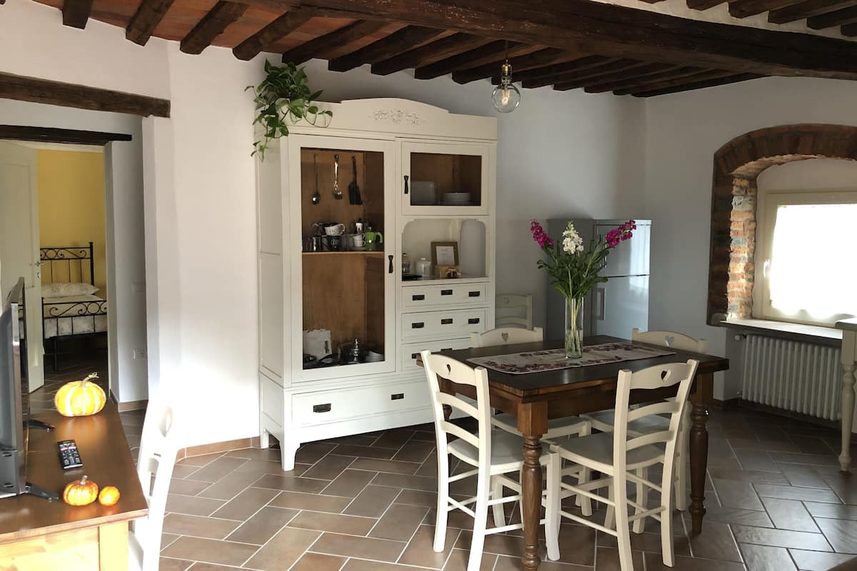 A dining area with a wooden table, white chairs, a white cabinet with glass doors, and a brick oven.