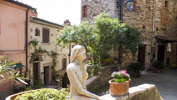 A stone building with a statue of a seated figure, potted plants, and a cobblestone street. - Le Dame del Borgo (Sassetta)