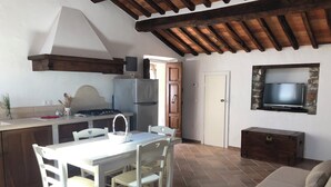 A compact kitchen with a dining area, a TV mounted on a stone wall, and a wooden ceiling. - Le Dame del Borgo (Sassetta)