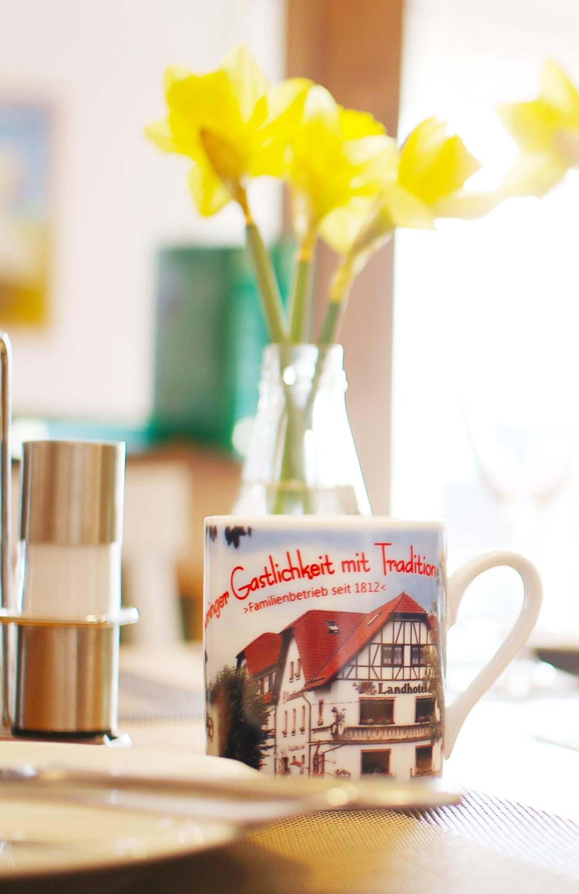 A mug with a traditional building illustration and text, a vase with yellow flowers, and a metal container on a table.