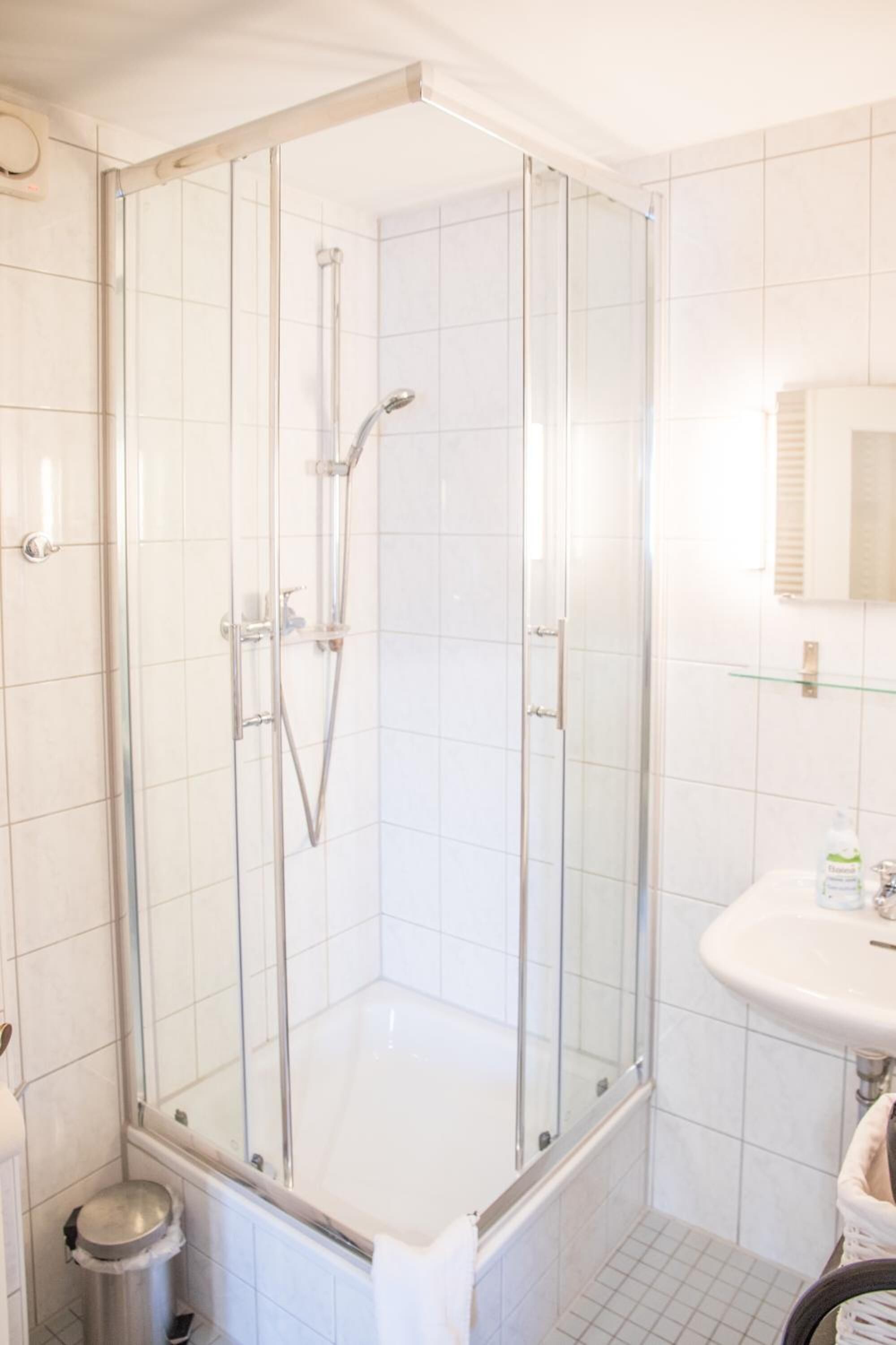 Ein modernes Badezimmer mit einer Duschkabine, weißen Fliesen, einem Waschbecken und einem Mülleimer.