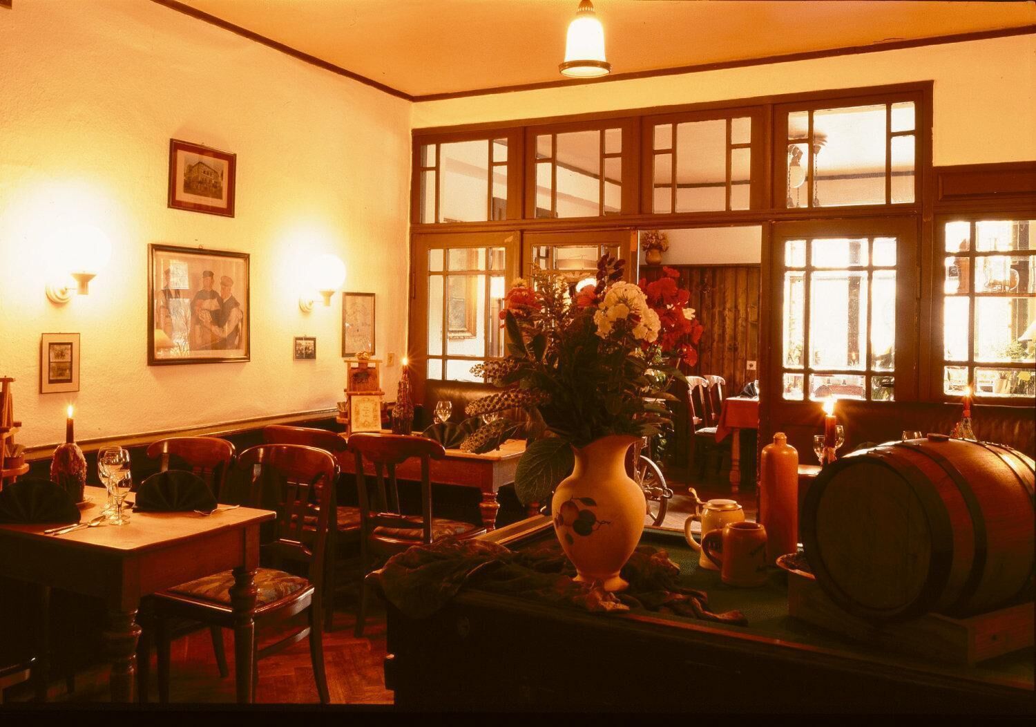 A dimly lit restaurant with wooden furniture, large windows, and a vase of flowers on the table.