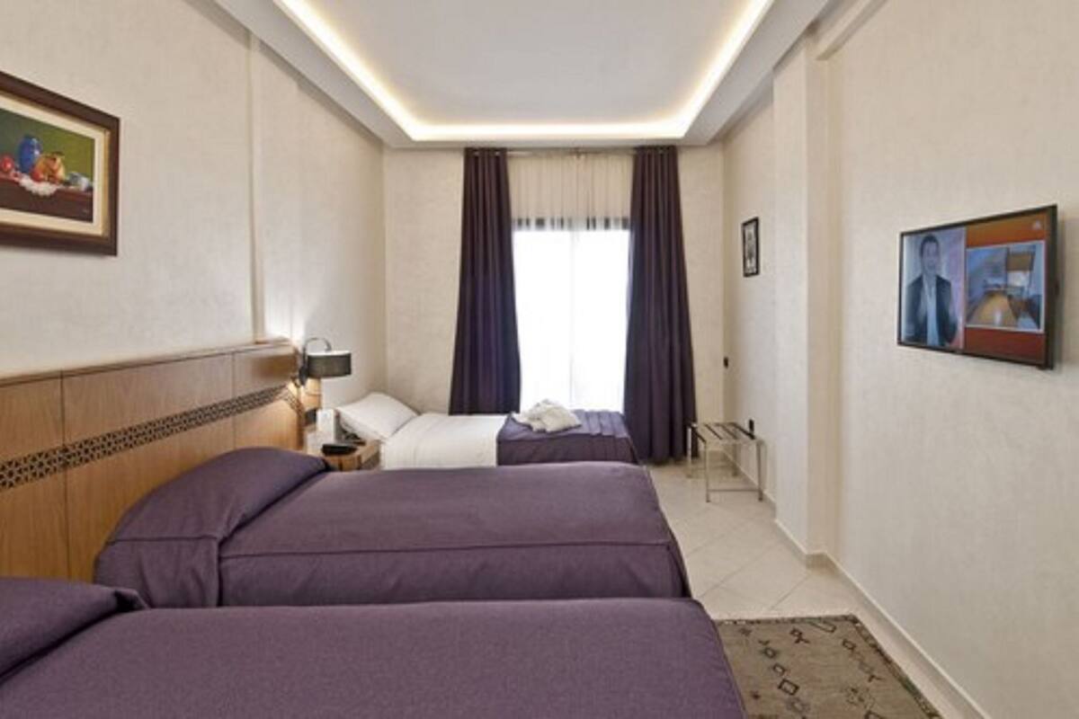 A hotel room with two beds, a nightstand, a lamp, a window with curtains, and artwork on the walls.