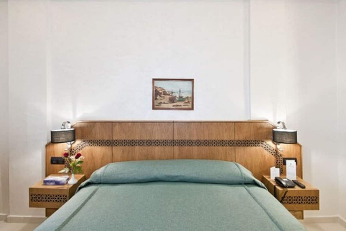 A hotel room with a wooden headboard, a green bedspread, a framed picture on the wall, and two nightstands with lamps and a phone.