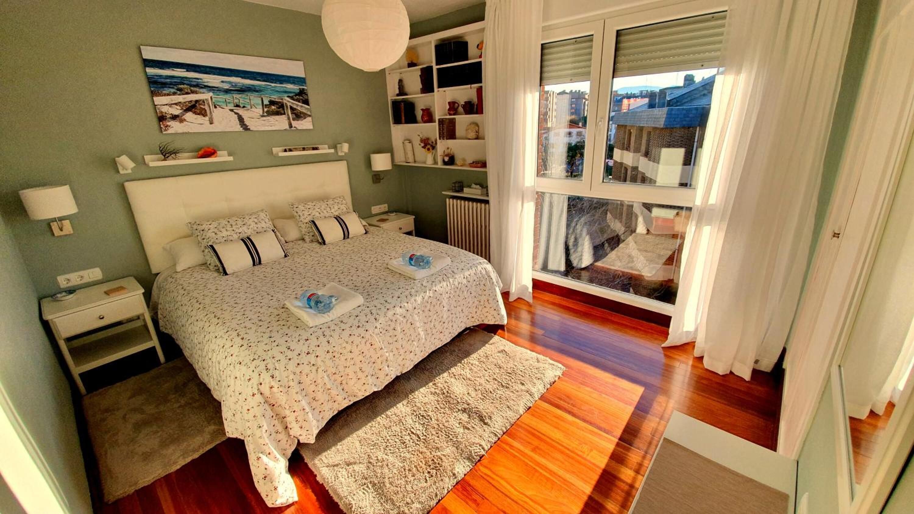 A bedroom with a bed, bedside tables, a nightstand, a window with a view, and a shelf with decorative items.