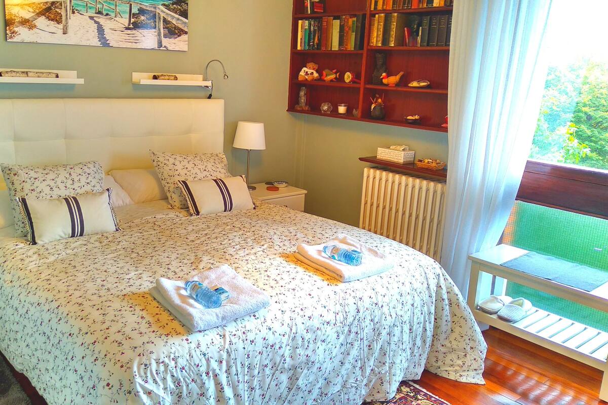 Un dormitorio con una cama grande, una mesita de noche, una estantería y una ventana que ofrece vistas al verdor.
