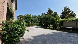 Une allée de gravier mène à un bâtiment à la façade en briques, entouré d’une végétation luxuriante et d’arbres.