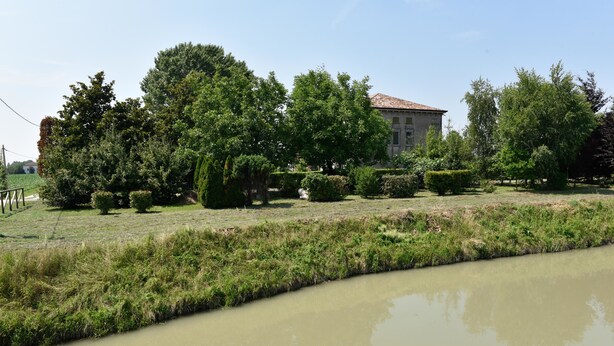 Une vue paisible sur la rivière, avec un bâtiment en arrière-plan, entouré d’une végétation luxuriante et d’arbres.