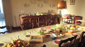 Una mesa de comedor dispuesta con frutas, velas y varios platos, rodeada de sillas de madera en una habitación con mantel estampado y una lámpara.