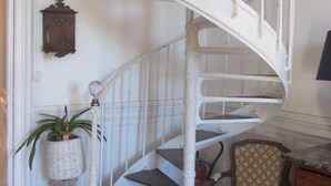 Una escalera espiral blanca con detalles de madera, una planta en maceta y una silla estampada en una habitación con paredes blancas y suelo de madera.
