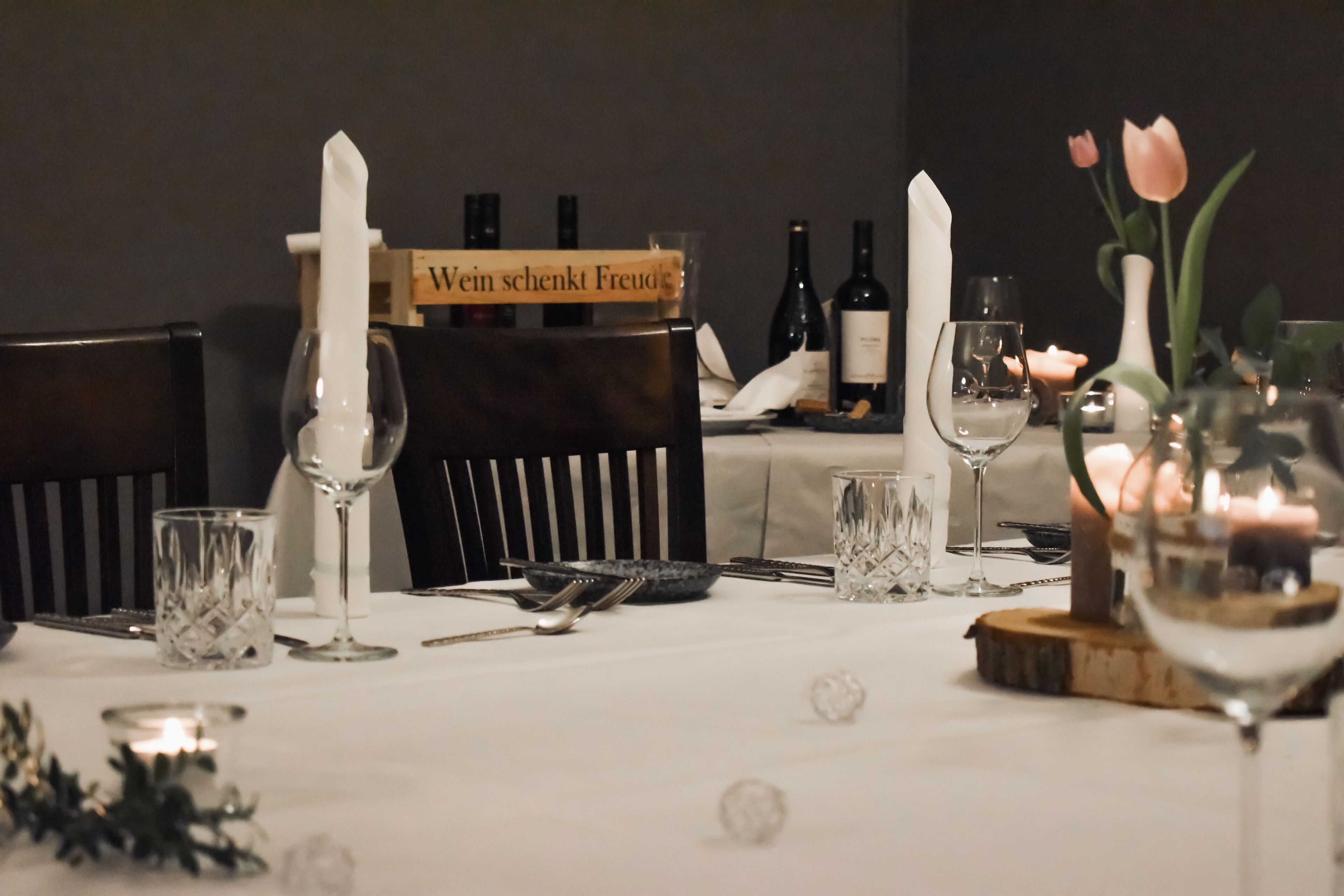 A dining table set with wine glasses, cutlery, and candles, with a sign reading "Wein schönkt Freud".