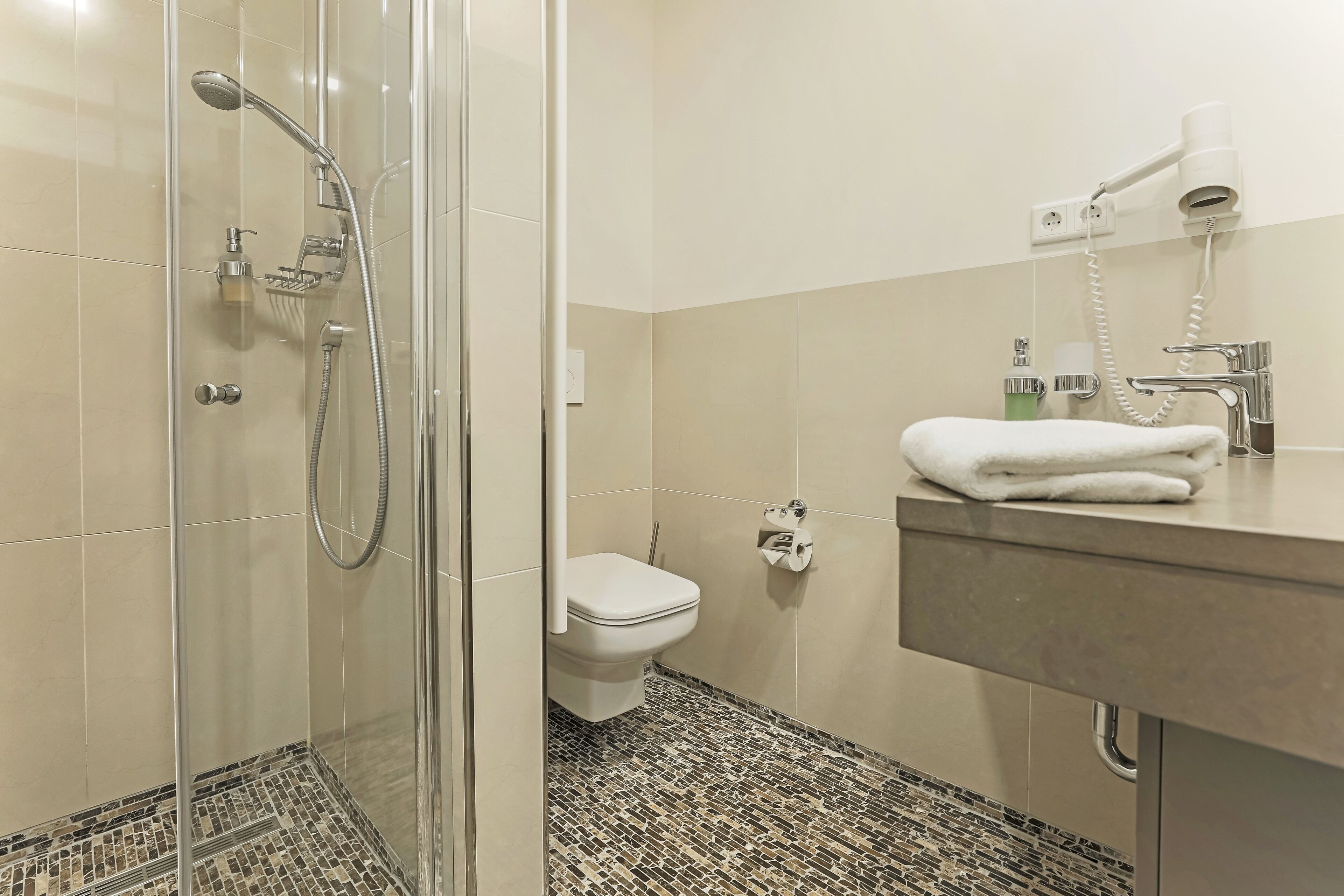 A bathroom with a glass-enclosed shower, a toilet, a sink with a towel, and a hairdryer on the wall.
