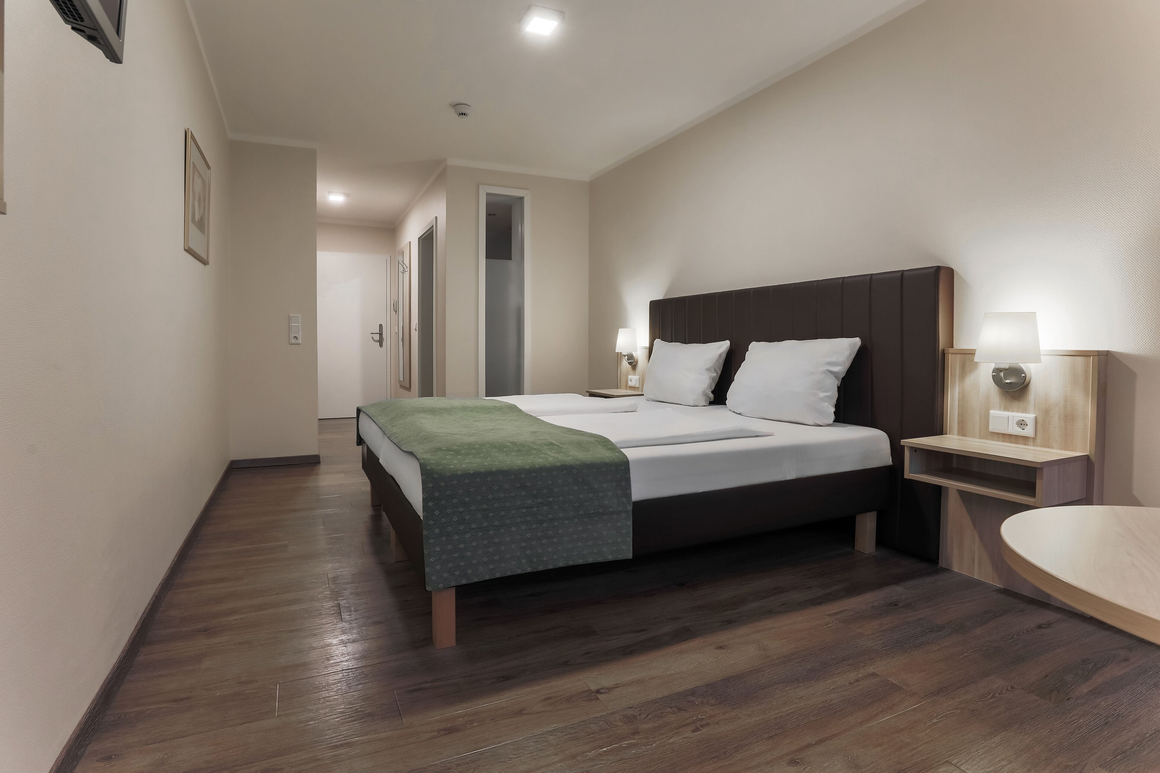A modern hotel room with a large bed, bedside tables, a wooden floor, and a hallway leading to other rooms.