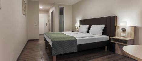 A modern hotel room with a large bed, bedside tables, a wooden floor, and a hallway leading to other rooms.