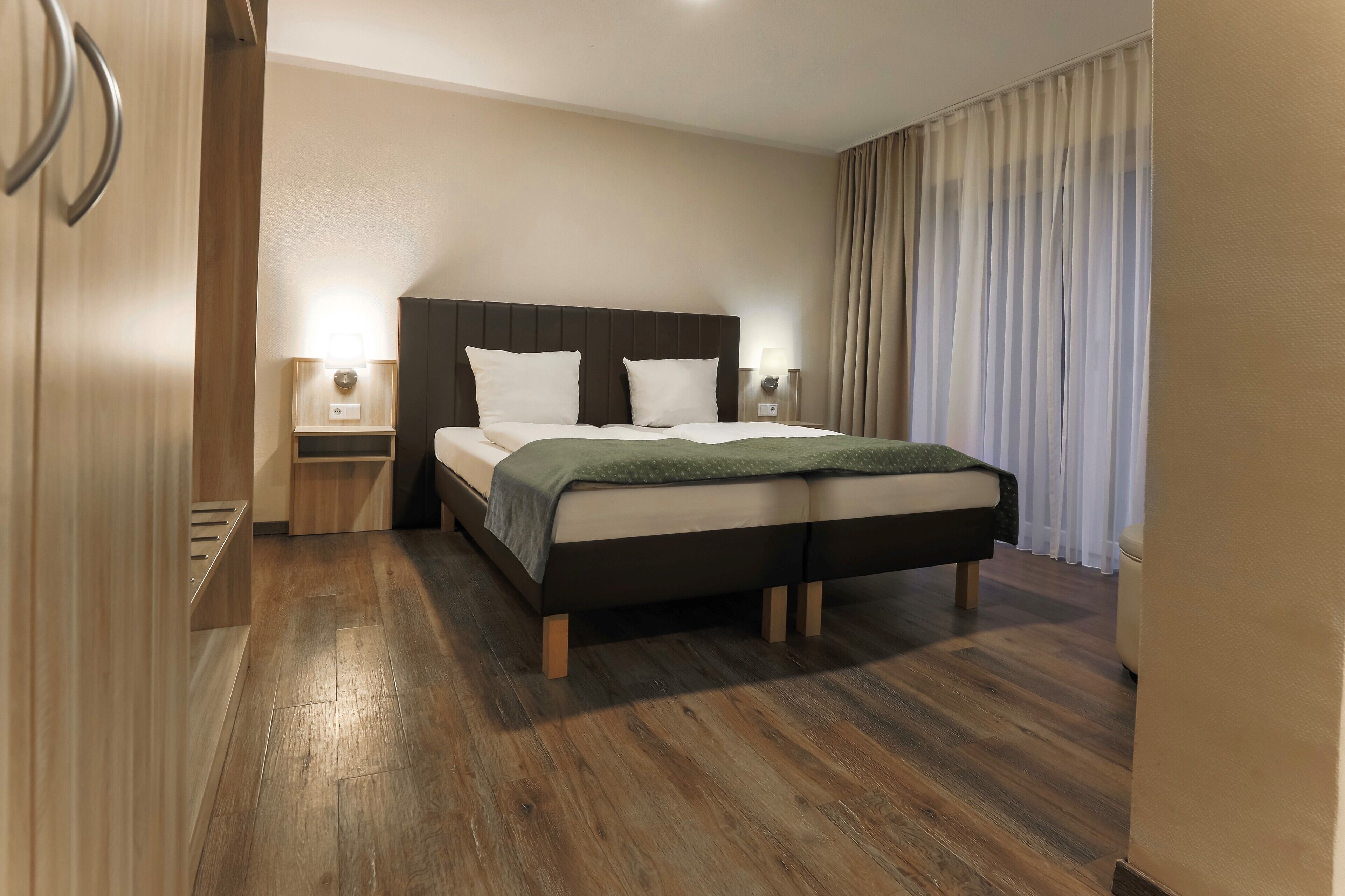 A hotel room with a large bed, wooden flooring, a headboard, two bedside tables with lamps, and a window with curtains.