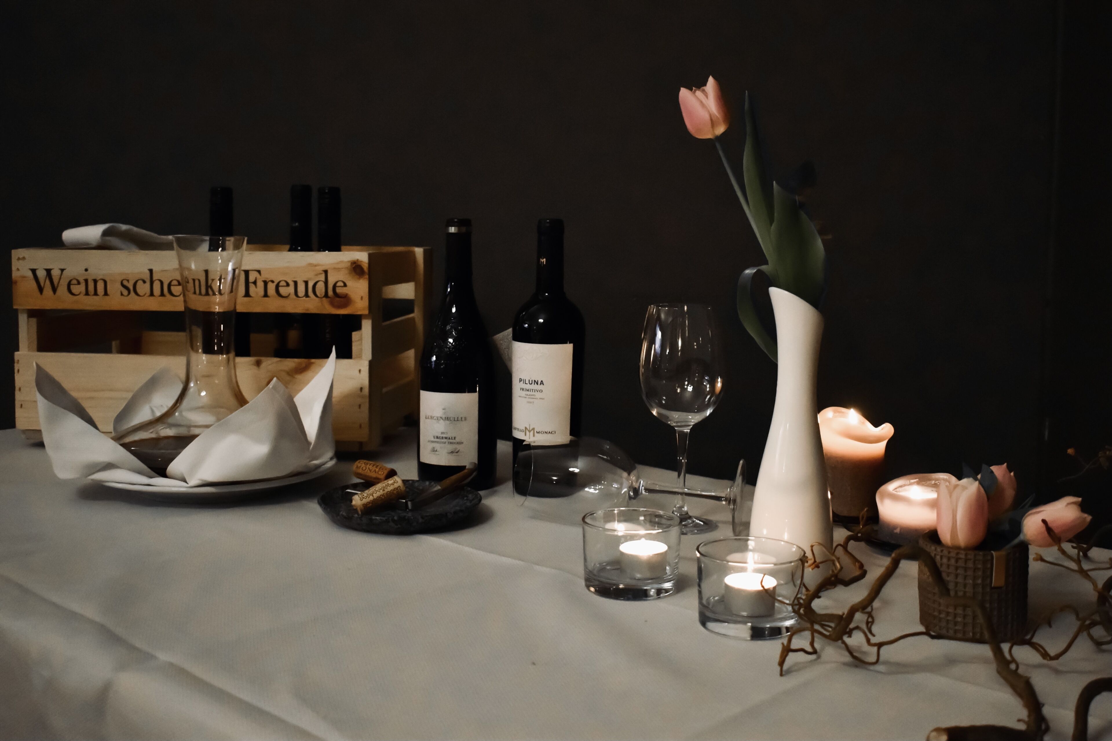 Una mesa con botellas de vino, copas, una vela y una caja de madera con la etiqueta 'Wein schekt Freude'.