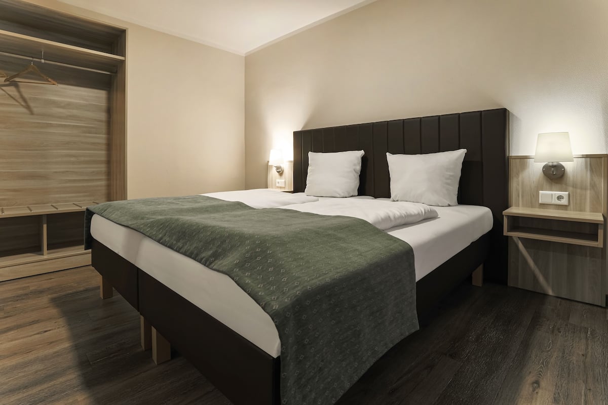 A modern hotel room with a large bed, wooden flooring, a wardrobe with a wooden door, and a bedside table with a lamp.