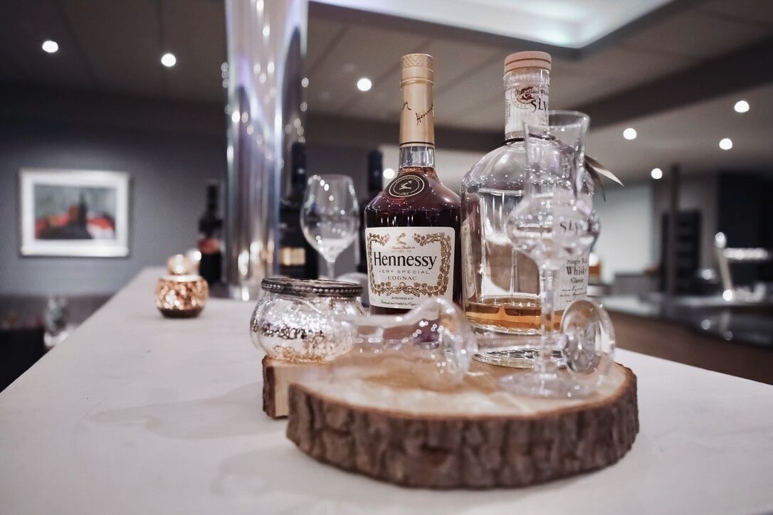 Una zona de bar con botellas de Cognac Hennessy y otras licores, copas y objetos decorativos sobre una superficie de madera.