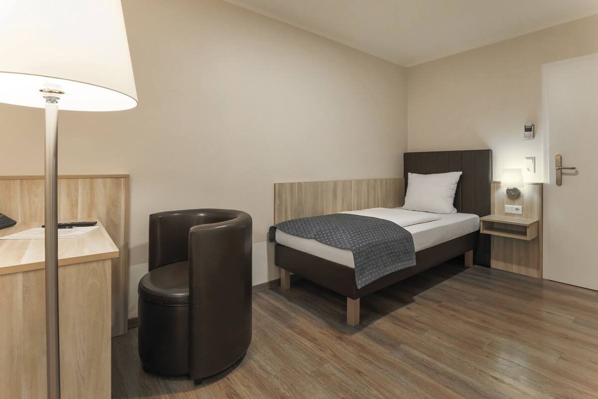 A modern hotel room with a wooden floor, a bed with a white and dark patterned blanket, a desk with a lamp, and a trash bin.